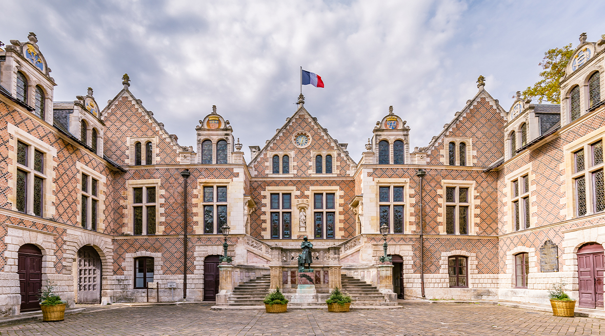 An impressive many gabled period building with its own courtyard, a French flag flies from the central gable and a statue stands in the middle of a set of steps